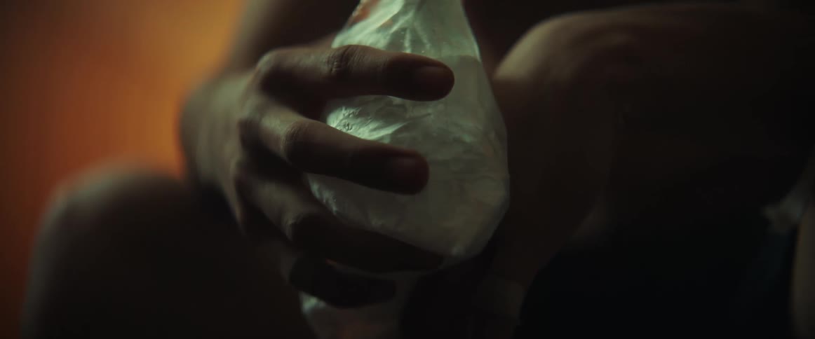 Young shirtless boxer sitting on a dimly lit room applying an ice pack over an injured hand