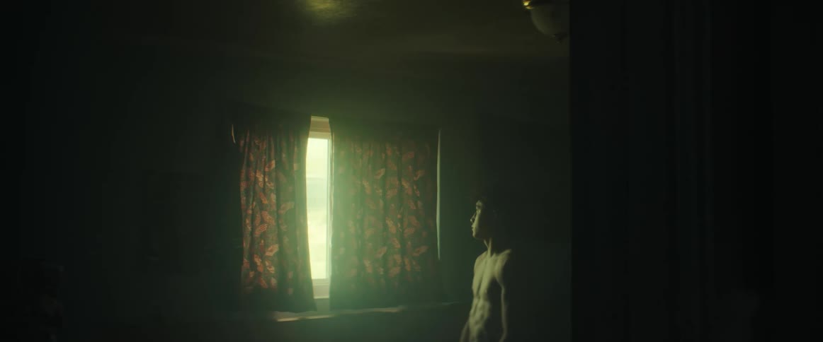 A teenage boy at home looks out a window and then reaches down and picks up a shirt and puts it on