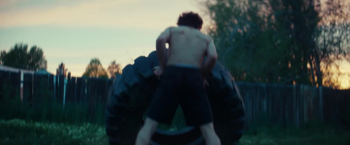 Muscular boy wearing black shorts pushes tire in the garden at sunset