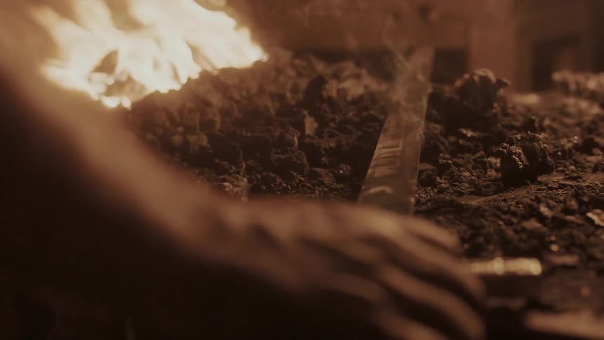 A man laying a sword on a pile of coals next to a fire