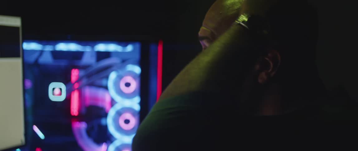 A man is sitting in a dimly lit room near a computer as he rubs his head with his hand