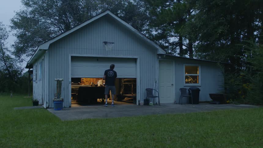 A man approaches a garage and lifts open the door before entering to retrieve a surfboard