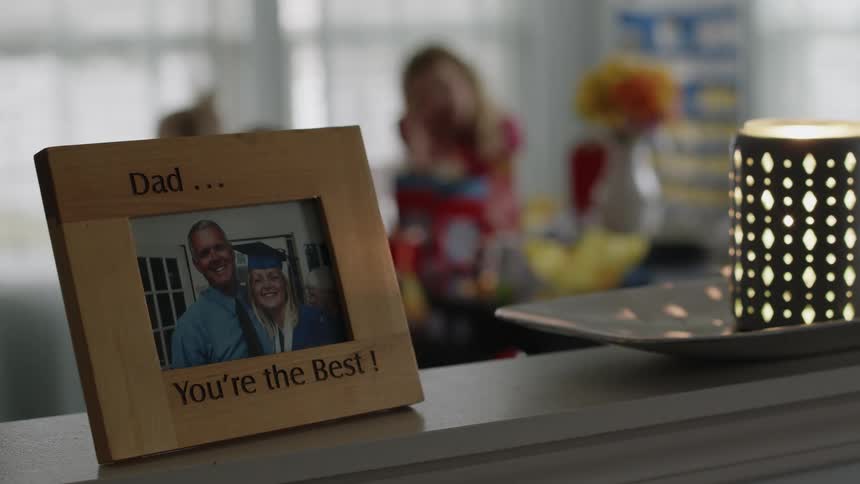 A photograph of a man and his daughter at her graduation is in a wooden frame on a table as people are sitting at a table in the distance