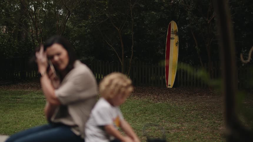 Daughter eats and mother talks on a cell phone in the park while a surfboard stands leaning against a fence in the background