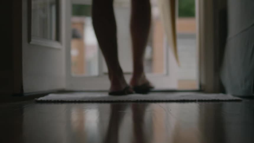 Barefoot woman puts on sandals and picks up a surfboard before getting out of the house