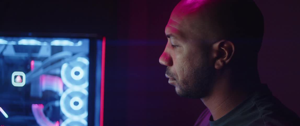 A man is sitting in a dimly lit room as the lights on his computer move and glow