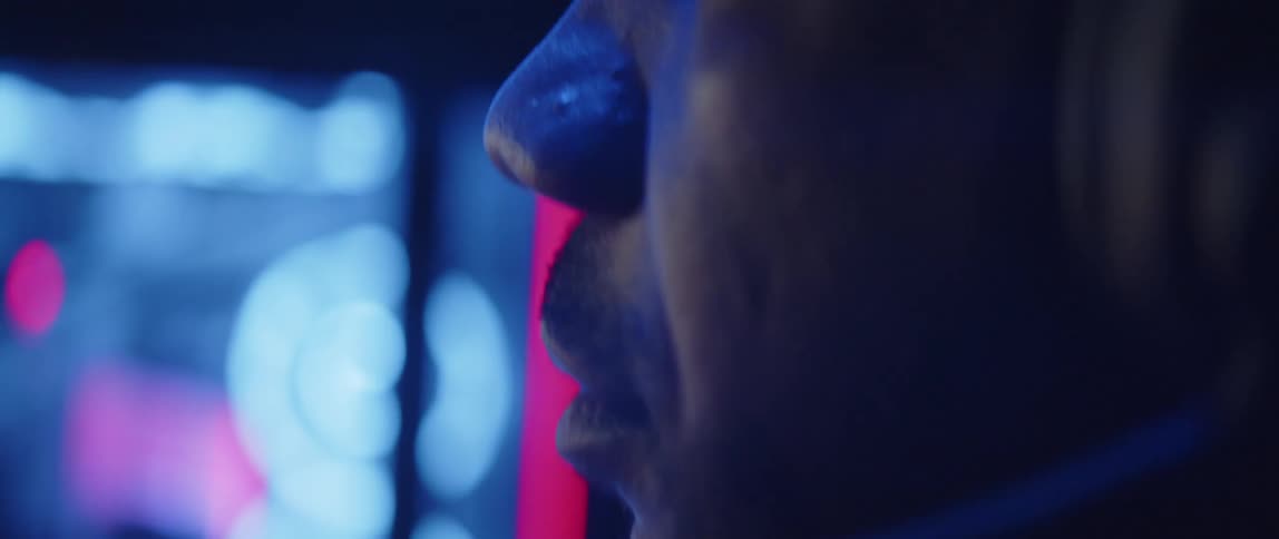 Man wearing headset sits at desk with large monitor in dim room lit by colored lighting