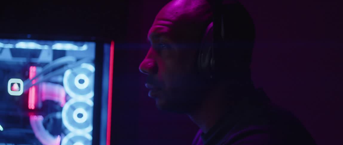 Bald man with silver watch wears headphones next to computer in dark room