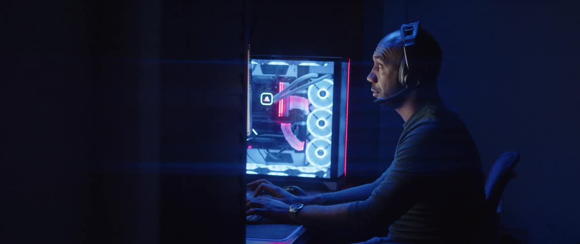 A man sitting at a desk types on a computer in a dark room illuminated by the computers lighting