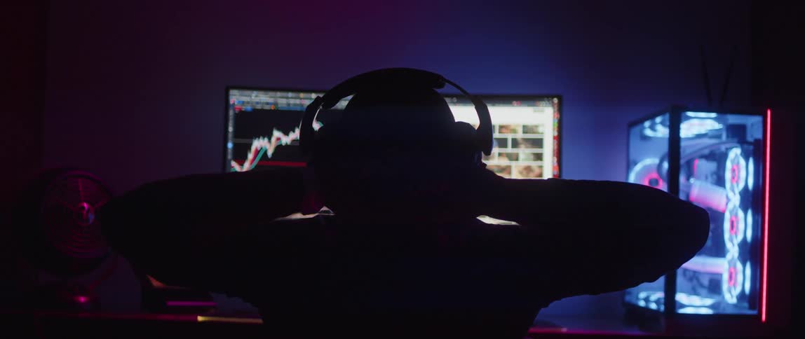 Silhouette of a stock trader wearing headphones with his hands on his head and a computer monitor in front of him and his computer on a table