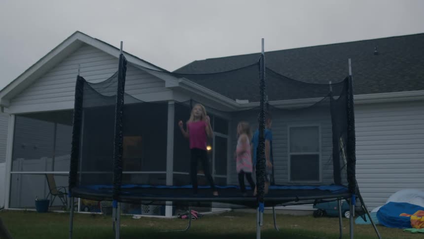 A little boy climbs up and joins his sister jumping on a trampoline in the back yard