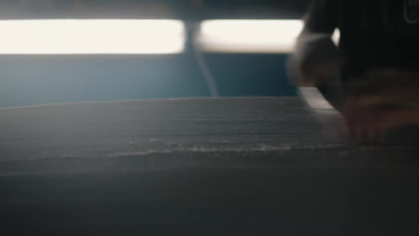 Man uses a tool to sand the surface of a surfboard inside a small workshop