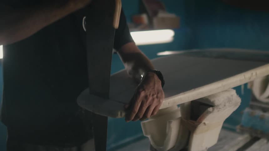Man with black t shirt and wristwatch cuts the top off of surfboard using handsaw