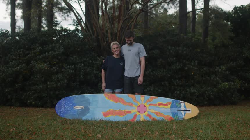 A man and a woman are standing behind a surfboard on the grass with their arms around each other with woods behind them