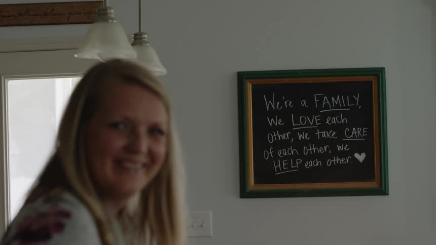 A woman is standing inside a room in her house looking at a hand written sign on her wall and laughing
