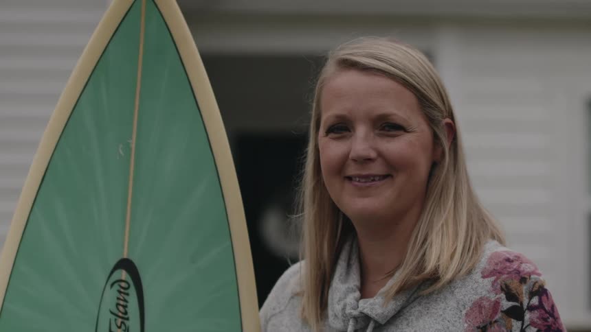 Happy blonde woman holding a surfboard in front of a house turns to look at the camera smiling