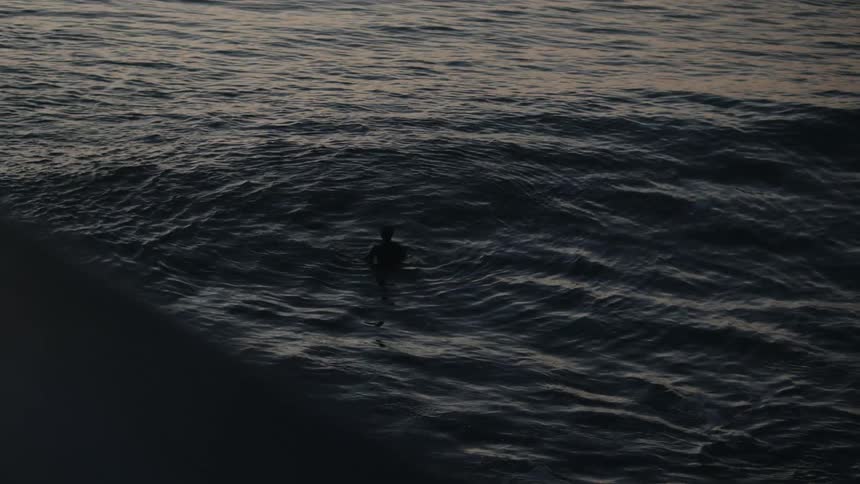 Man silhouetted in dim light bobs in water as waves roll past him