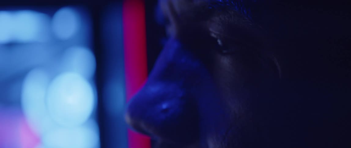 A man sits in a dimly lit room as lights flash on his face and lights glow next to him