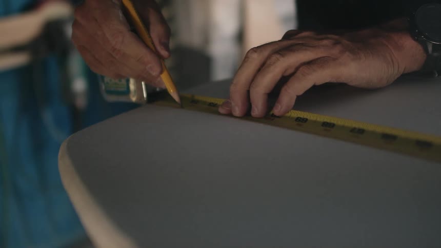Close up of a man crafting a surfboard making a mark with a pencil using a measuring tape