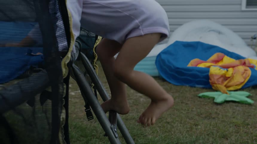 Blonde toddler girl gets on trampoline to jump in the garden next to building