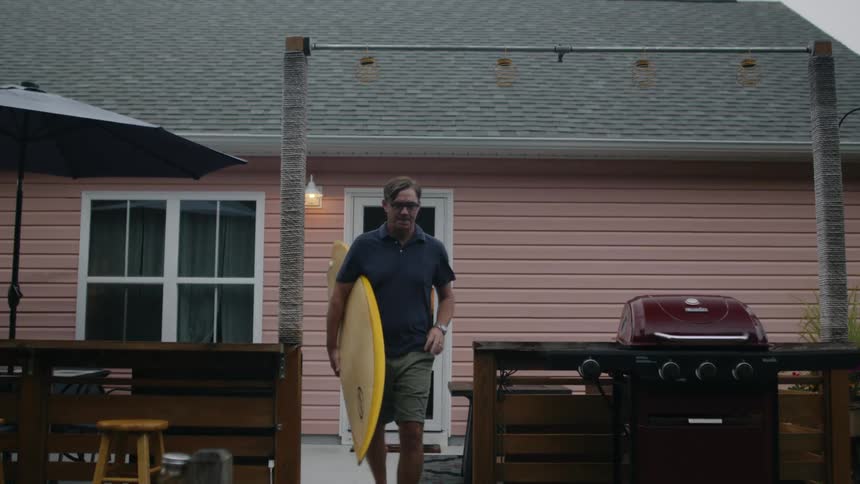 A man carries a surfboard out of a house and walks out of frame