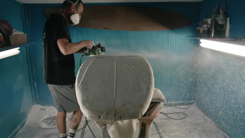 Man crafting a surfboard uses a power tool to sand the surface inside a small workshop