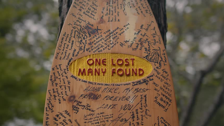 Surfboard shaped wooden plaque hangs on tree in remembrance with many messages written on it