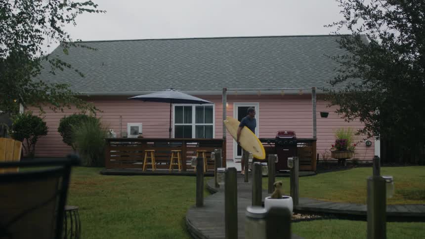 A man carries a surfboard under his arm while leaving his house