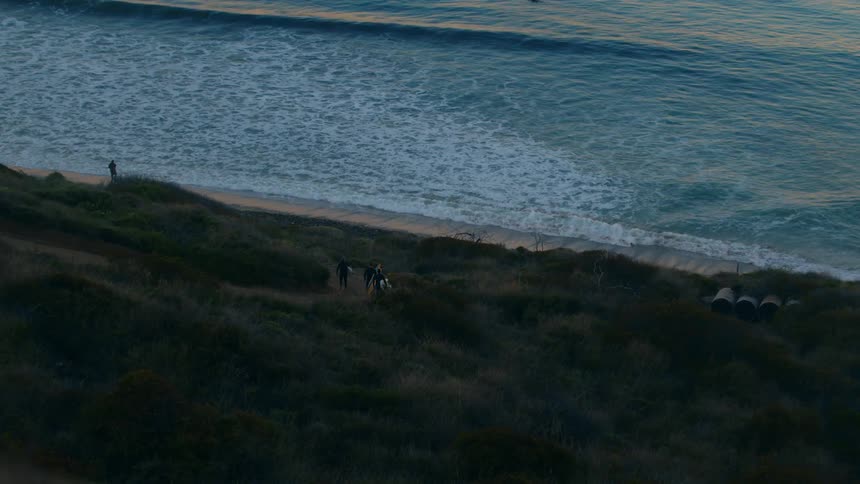 A group of surfers carry their surfboards down a slope towards the beach below