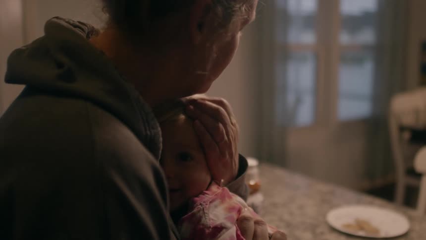 Mother with gray hoodie hugs daughter with pink and white top next to dining table in the kithcen