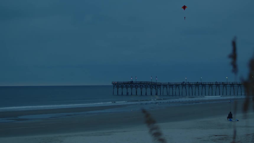 Man flies kite on the beach near the ocean and long pier at dusk