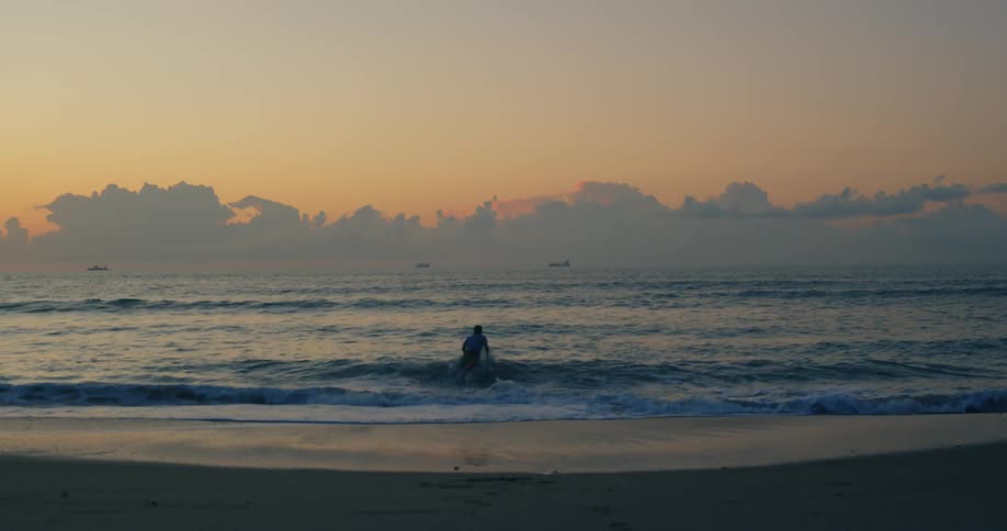 Male surfer going surfing and runner jogging on the beach are silhouetted against the sky at dusk