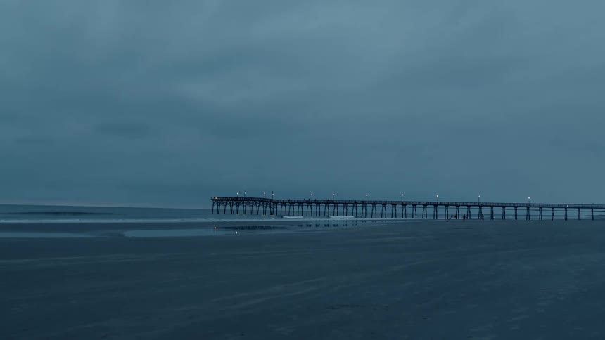 The silhouette of people playing and walking near the pier on an overcast gray day at dusk