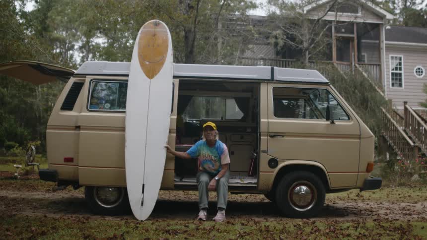 A woman sitting in the open door of her van smiling and looking at her surfboard