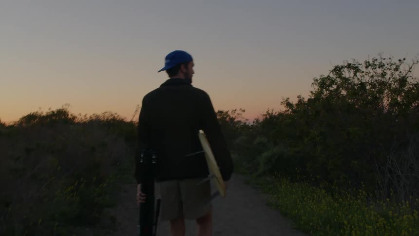 Man with blue cap black sweater and gray shorts walks on track in the field carrying surfboard and tripod at sunset