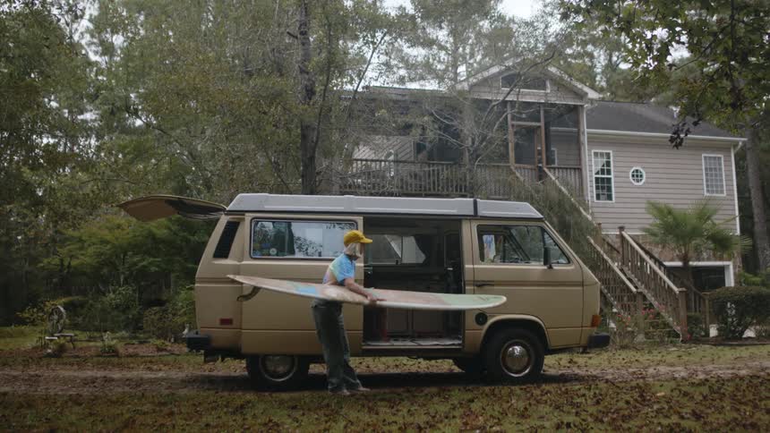 Woman with yellow cap blue white t shirt and gray pants takes surfboard out of van