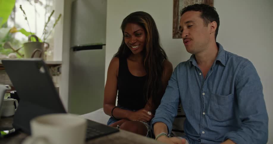 A man and a woman are sitting down together looking at a laptop computer on a table and talking and laughing