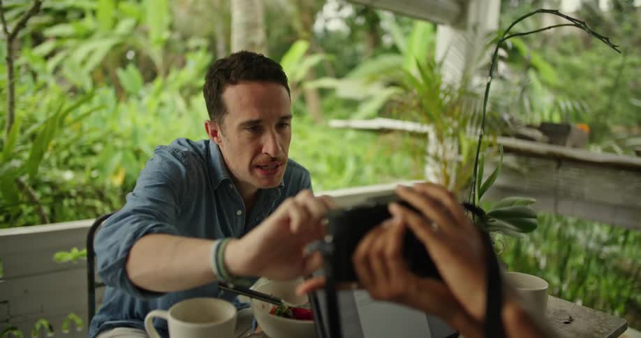 A woman holds a camera and shows it to a man across the table from her and he talks and pushes some buttons