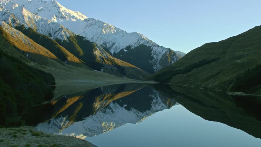 Aerial view of the calm surface of a lake perfectly reflecting the snow of a mountain range at dawn
