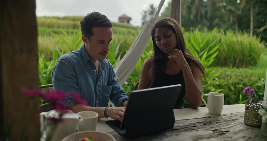 A man and woman sit at a table outside and he types on a laptop as she watches