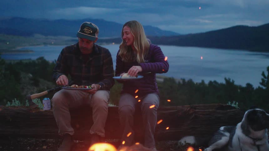 Man with cap plaid shirt and brown pants sits next to blonde woman with purple jacket and jeans by campfire with lake and mountains in the background