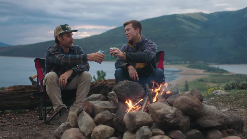 Two men talk and have drinks by the campfire