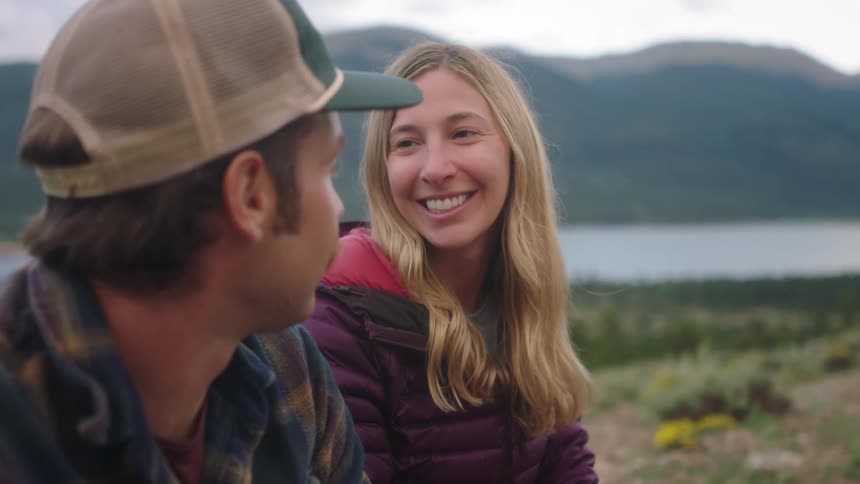 Man with cap mustache and blue shirt talks with blonde woman wearing purple coat holding a can of beer laughing and sitting on hill overlooking lake and high mountains
