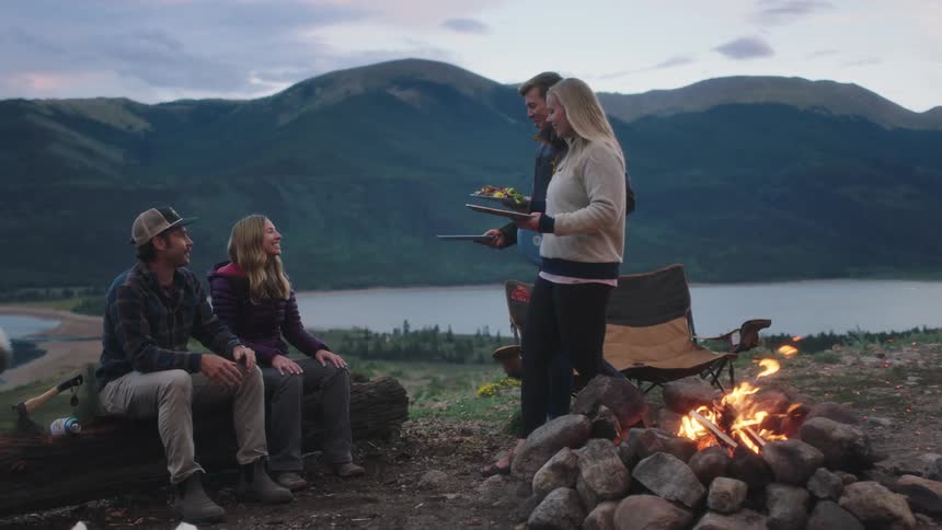 Couples sharing food next to campfire camp on hill overlooking high mountains and lake