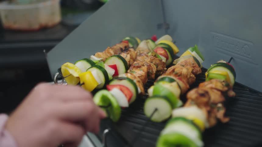 A person turning skewered meat and vegetables cooking on a grill