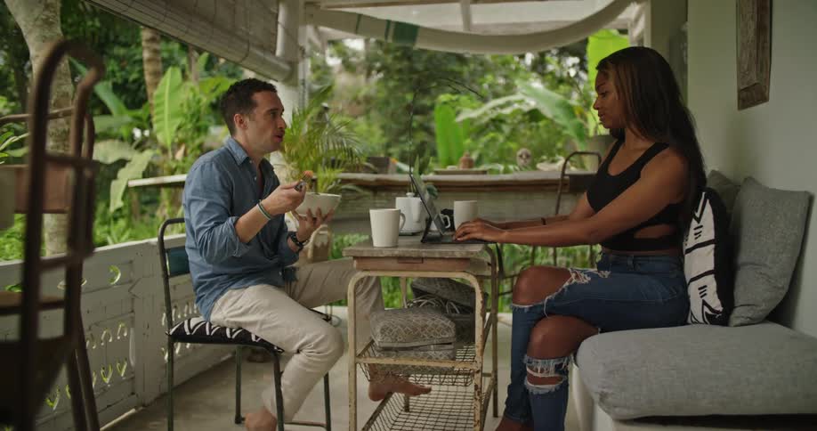 A woman types on her laptop as a man eats fruit from a bowl while they sit on their patio