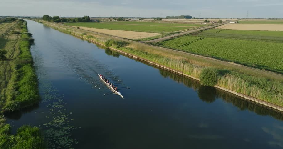 Aerial view of a rowing team on a racing boat over a river next to the farming fields