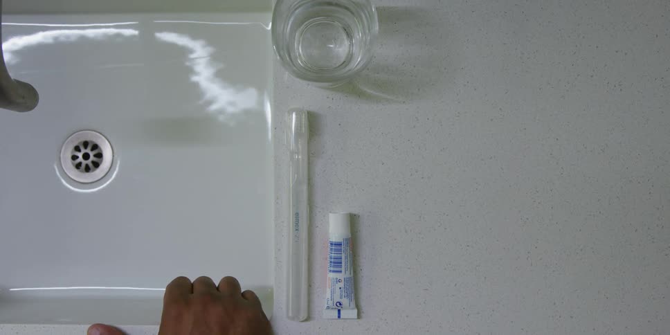 A person puts their hand on a sink next to a small tube of toothpaste and a glass