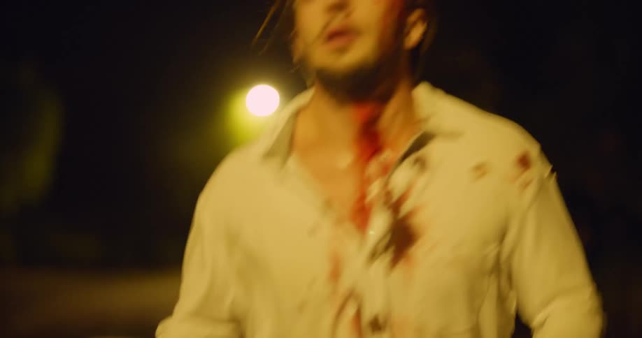 Injured man wearing a white shirt stained with blood runs and collapses on an empty road at night