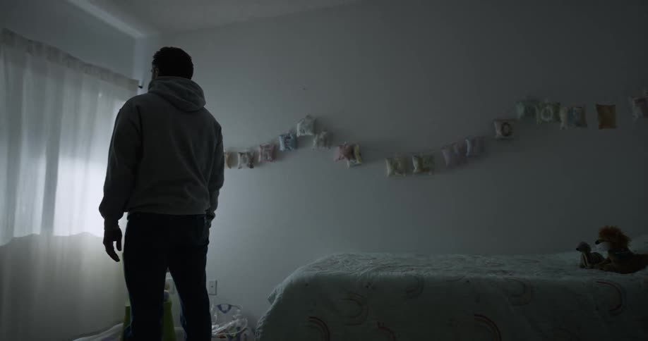 Man with gray hoodie and jeans stands next to bed in room